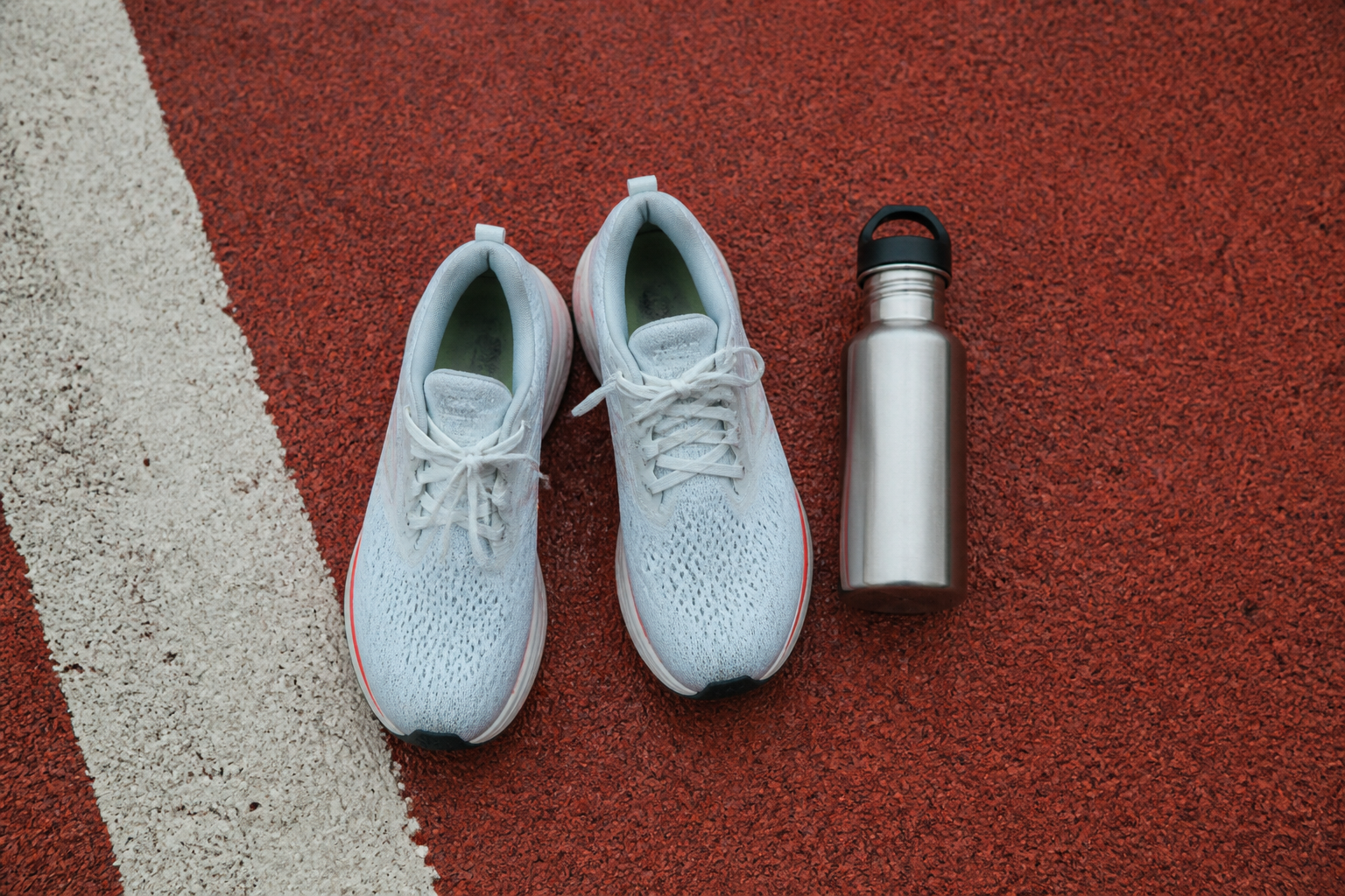 A top-down view of a pair of high-end running shoes and a reusable water bottle on a running track, soft overcast lighting.