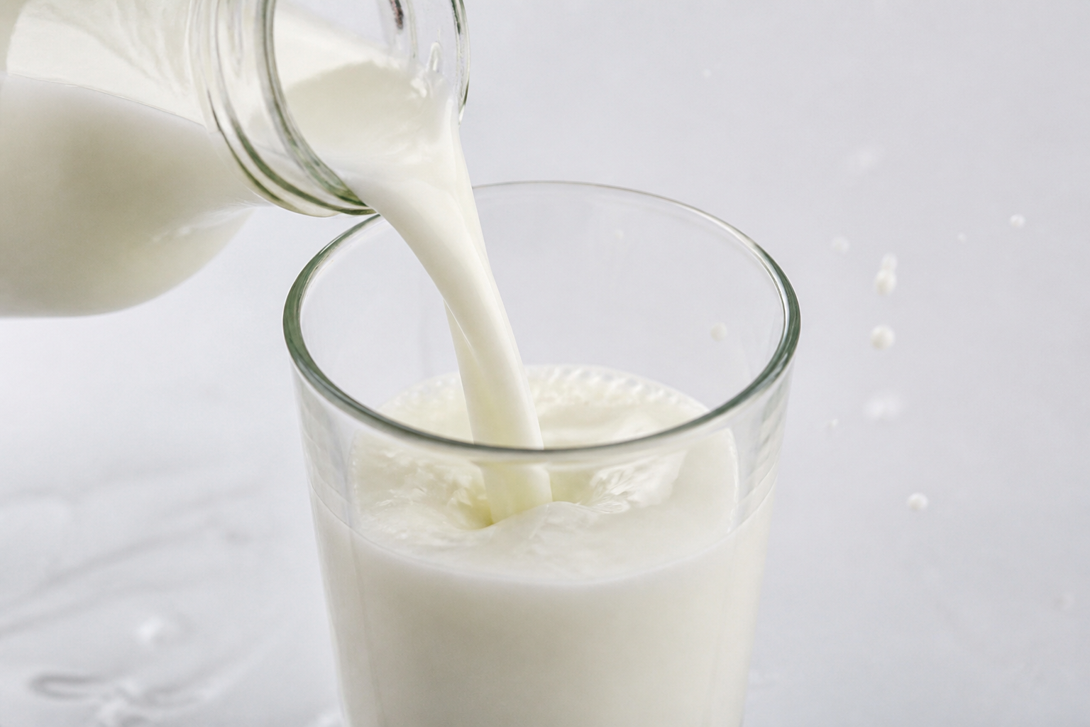 A close-up shot of fresh white milk being poured into a clear glass, with small droplets and a clean white background.