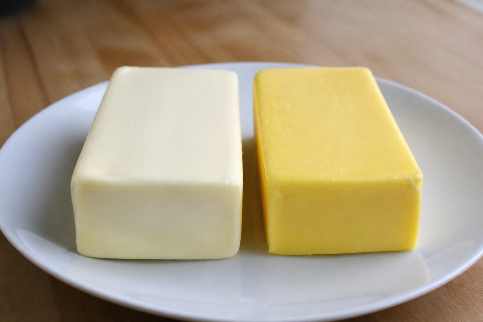 Two blocks of butter side-by-side on a white plate: one pale white from indoor cows, one deep yellow from pasture-raised cows.