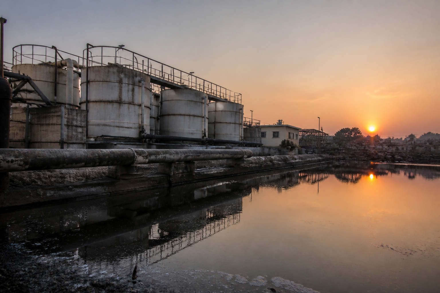 Large industrial filtration tanks and pipes of a water treatment facility at sunset, reflecting off a nearby still pond.