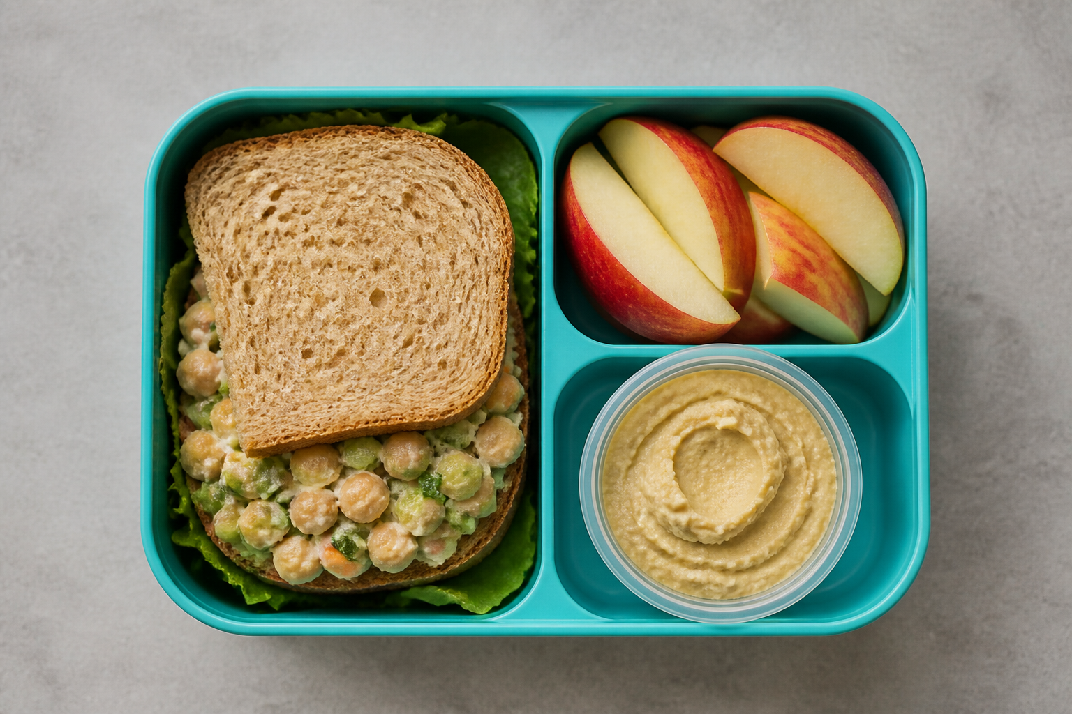 A top-down view of a colorful school lunch box containing a chickpea salad sandwich, sliced apples, and a small container of hummus, neutral background.