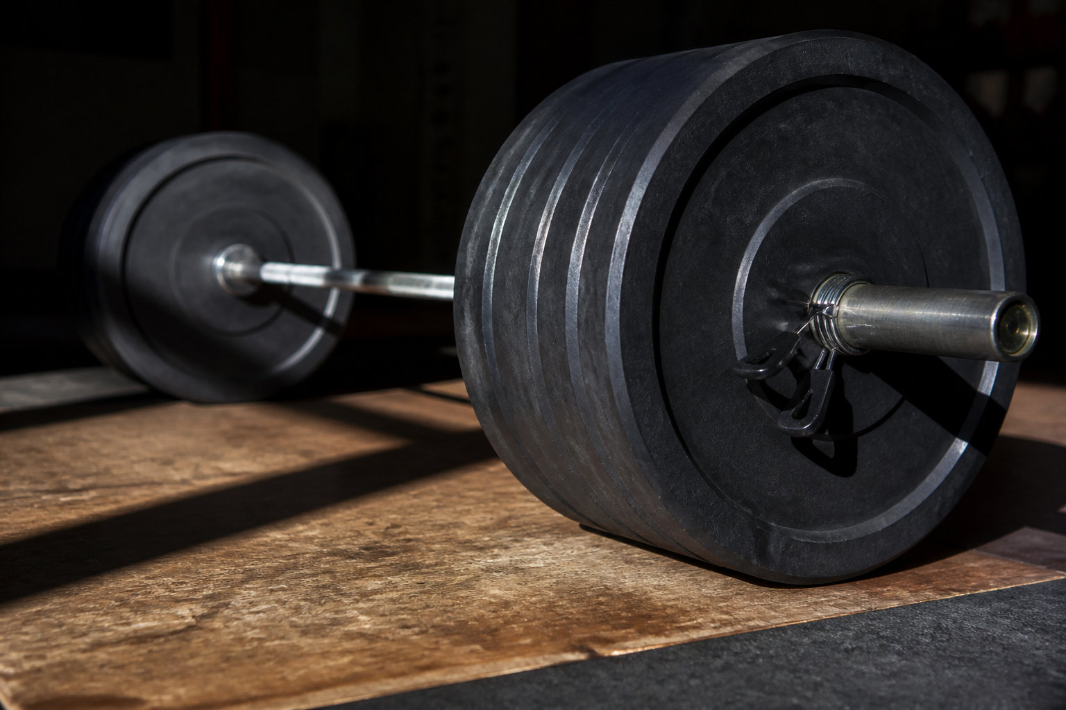 A close-up of a professional Olympic barbell loaded with multiple heavy black plates on a wooden lifting platform, dramatic side lighting.