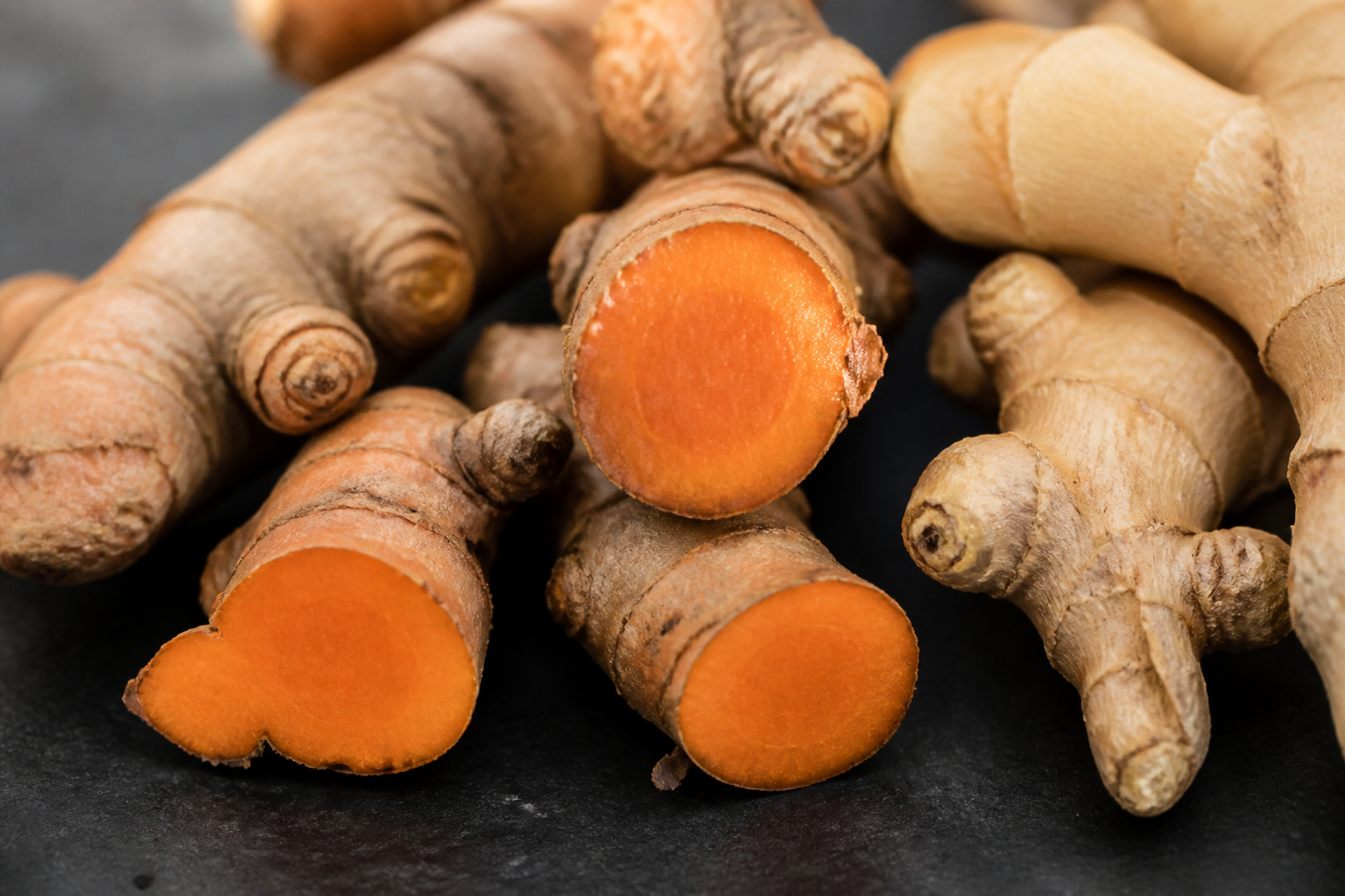 Macro photograph of fresh turmeric and ginger roots on a dark slate background, showing the textured skin and bright orange interior of a sliced turmeric root.