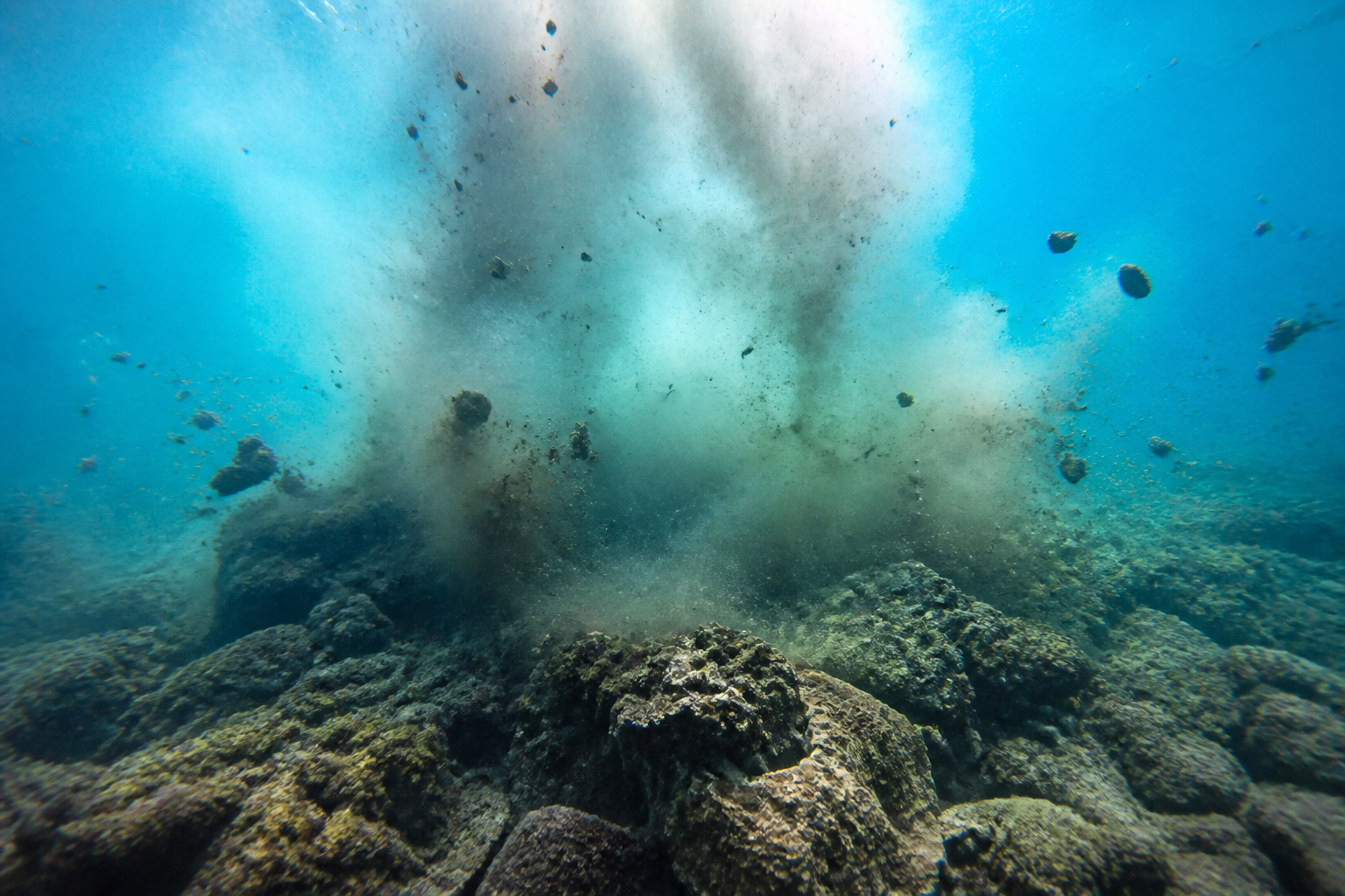 A dramatic underwater photograph capturing the moment of a distant explosion, with air bubbles and debris clouding the turquoise water.